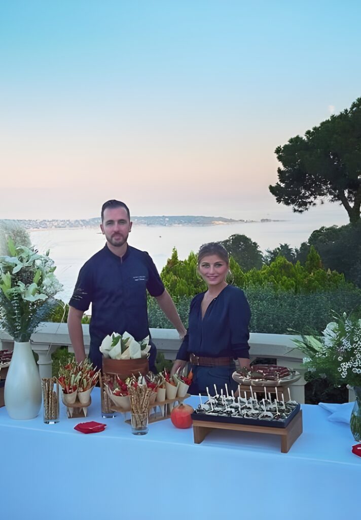 Buffet dînatoire chic face à la mer au crépuscule Chef et femme souriants derrière une table de service extérieure avec apéritifs et fleurs, devant un paysage marin au coucher du soleil.