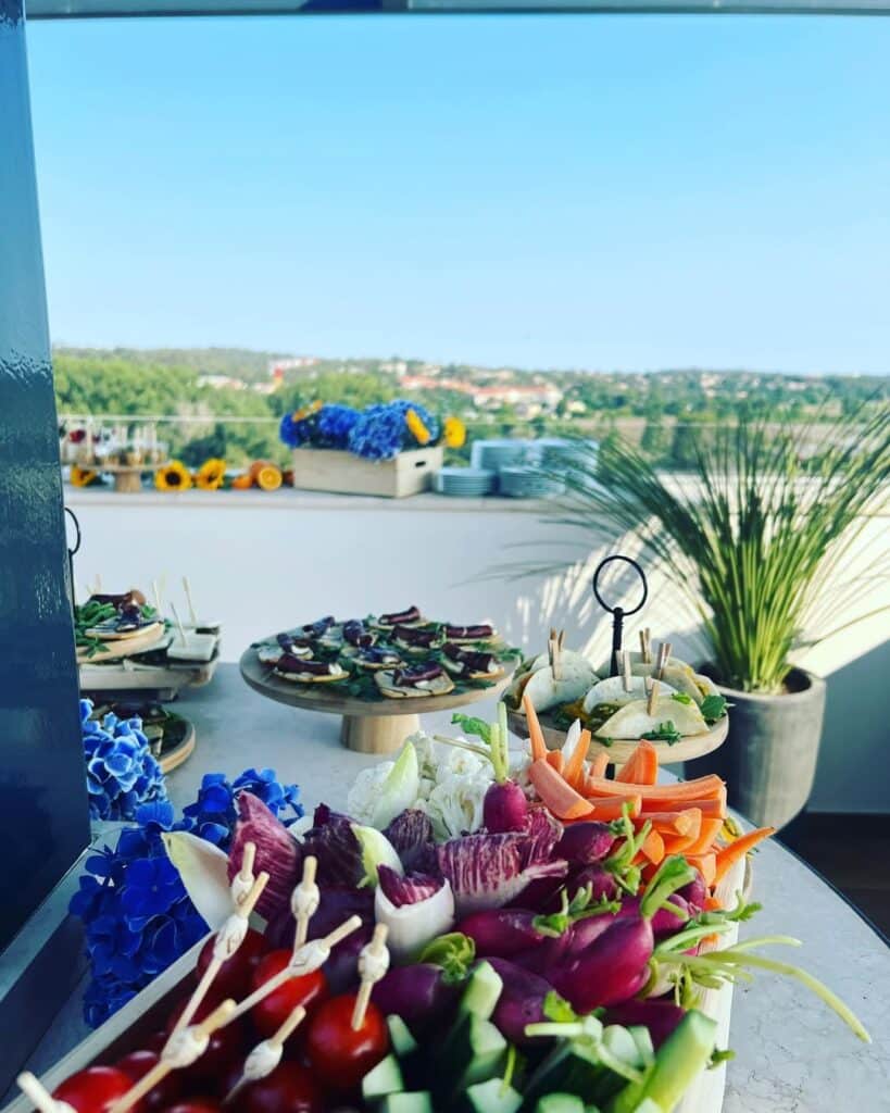 Buffet Dînatoire Extérieur Vue Panoramique et Raffinée Buffet extérieur avec plateau de crudités fraîches (radis, carottes, concombres) et hors-d'œuvre sur une terrasse ensoleillée avec vue sur une ville verdoyante.