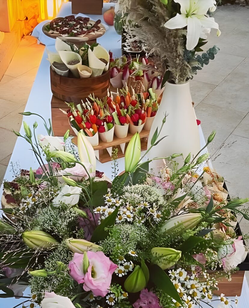 Buffet Traiteur Élégant avec Fleurs Fraîches Buffet extérieur élégant avec bouquets de lys blancs et roses, légumes coupés en brochettes et charcuteries sur table blanche.