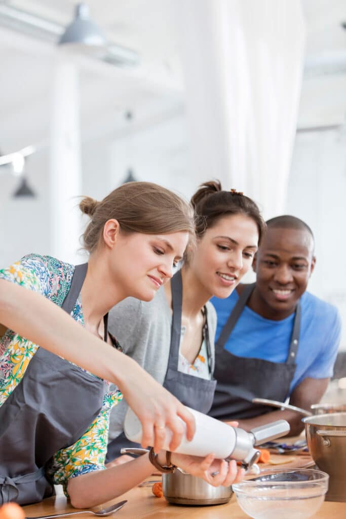Cours de cuisine : amis préparant des recettes ensemble Trois adultes souriants, deux femmes et un homme, portent des tabliers et cuisinent ; une femme utilise un siphon à crème sur une casserole.