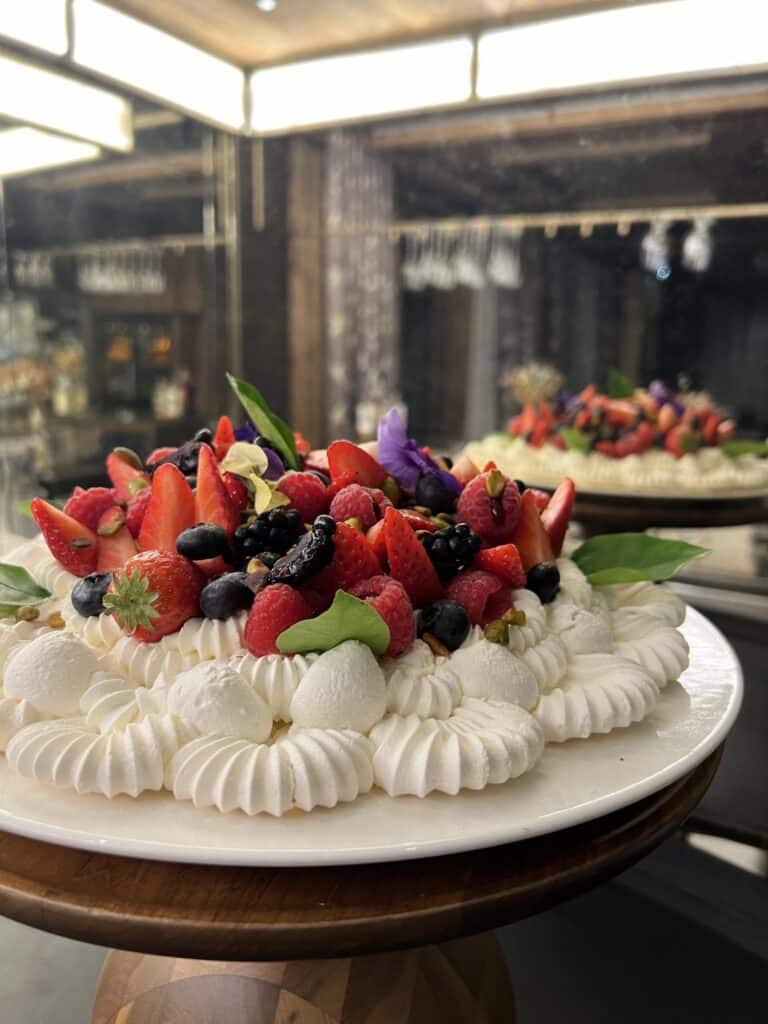 Pavlova aux Fruits Rouges : Délicatesse Meringuée Pavlova spectaculaire garnie de crème fouettée, fraises, framboises, myrtilles et fleurs comestibles sur une assiette blanche.